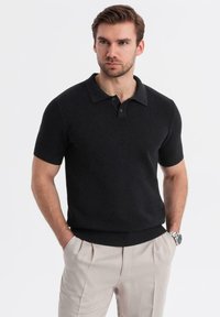 Camisa polo negra de manga corta con un diseño de punto texturizado, tapeta de tres botones y puños acanalados. Lleva puesta con pantalones de color claro.