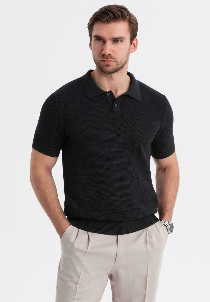 Camisa polo negra de manga corta con un diseño de punto texturizado, tapeta de tres botones y puños acanalados. Lleva puesta con pantalones de color claro.