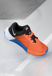 Sportssko med en lys orange overdel, sorte detaljer, blå sål, åndbart mesh-materiale og sorte snørebånd. Bemærkelsesværdige logoer.