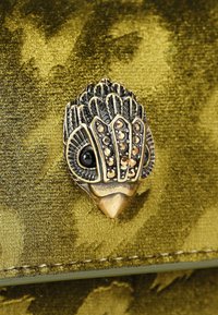 Sierlijke vogelgesp in bronskleur op een getextureerde olijfgroene stoffen achtergrond; gedetailleerd ontwerp met nep-edelstenen en een glanzend oog.