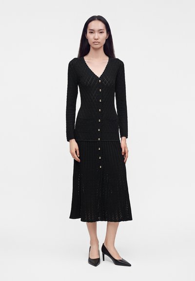 Femme debout sur un fond uni, portant une longue robe en tricot noire boutonnée, manches longues et talons pointus noirs.