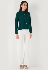 Türkise Bluse mit großen Kragen, kombiniert mit weißen ausgestellten Hosen und metallischen High Heels, vor einem neutralen Hintergrund stehend.
