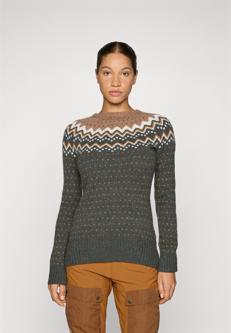 Knitted sweater in dark green with a textured pattern, featuring a light brown yoke and zigzag designs in white and orange. Long sleeves.