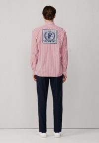 Rot-weißer gestreifter Langarmshirt mit einem blauen gestickten Logo auf dem Rücken. Kombiniert mit dunkelblauen Hosen und weißen Schuhen.