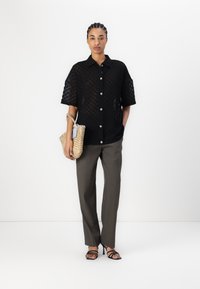 Chemise noire transparente à motif, pantalon gris ajusté, sandales à lanières noires et un sac en osier avec une longue anse.