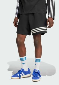 Pantaloni sportivi neri con strisce bianche, abbinati a scarpe da ginnastica in suede blu e calzini bianchi con strisce blu. Design semplice e sportivo.