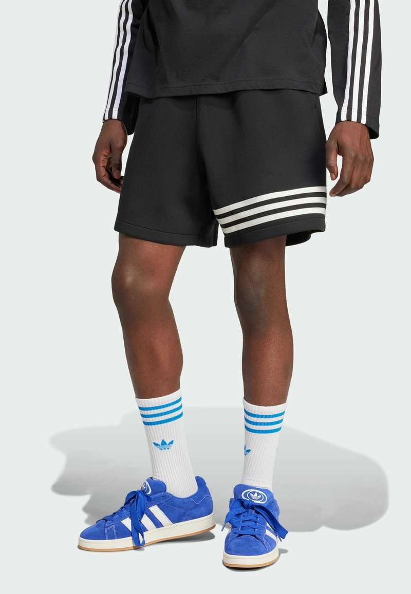 Pantaloni sportivi neri con strisce bianche, abbinati a scarpe da ginnastica in suede blu e calzini bianchi con strisce blu. Design semplice e sportivo.