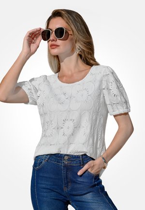 Kobieta w białej koronkowej bluzce w kwiaty i ciemnoniebieskich dżinsach, poprawiająca duże okulary przeciwsłoneczne jedną ręką, stojąca na jednolitym tle.