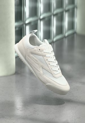 Witte Calvin Klein sneaker met mesh- en suèdetails, veters gestrikt, zwevend boven een gepolijste grijze vloer nabij een glazen blokmuur.