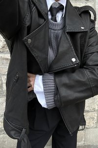 Schwarze Lederbikerjacke mit silbernem Zubehör, über einem grauen Strickpullover und einem weißen Kragenhemd kombiniert, das ein texturiertes Muster aufweist.
