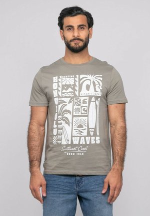 Homme barbu aux cheveux foncés portant un T-shirt gris avec des motifs de surf blancs et un jean bleu, sur un fond uni.