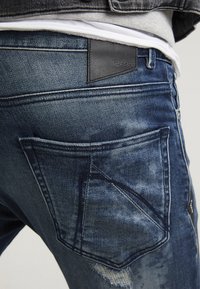 Jeans de denim azul con un acabado desgastado, que presentan una etiqueta de cuero oscuro en la cinturilla y detalles de costura únicos en los bolsillos.