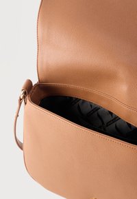 Καφέ δερμάτινη τσάντα με χρυσό κούμπωμα, ανοικτό καπάκι που δείχνει την εσωτερική επένδυση από μαύρο ύφασμα με το μοτίβο λογοτύπου Armani Exchange.