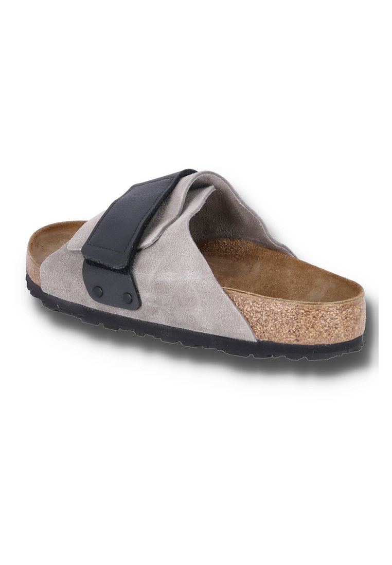 Graue Wildleder-Sandale mit Korkfußbett, schwarzem verstellbarem Riemen und schwarzer Gummisohle, in einem Winkel auf einem weißen Hintergrund dargestellt.