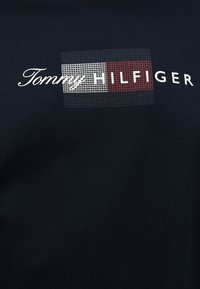 Tommy Hilfiger logo with white and red grid pattern on a dark fabric background.