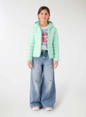 Ragazza giovane che indossa una giacca imbottita verde chiaro, una t-shirt bianca con grafica, jeans larghi strappati e scarpe da ginnastica rosa, in piedi davanti a uno sfondo bianco semplice.