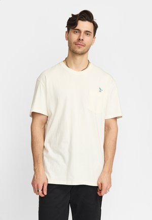 Uomo con capelli castani corti che indossa una maglietta a maniche corte color crema con taschino sul petto e piccolo logo di uccello colorato, in piedi davanti a uno sfondo uniforme.