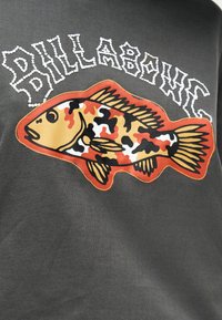 Έγχρωμο γραφικό ψαριού με μοτίβο παραλλαγής και αιχμηρό λευκό κείμενο «BILLABONG» σε σκουρόχρωμο γκρι φόντο ύφασματος.