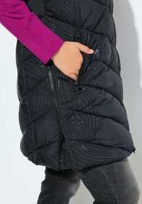 Ruka v zipové kapse černého vzorovaného prošívaného saka, které je oblečeno přes magentové dlouhé tričko a šedé kalhoty.