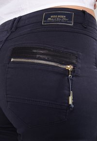 Los vaqueros azul marino cuentan con un pequeño bolsillo con cremallera con una superposición de malla negra, cremallera plateada y acentos de hardware dorado. Etiqueta MOS MOSH en la cinturilla.