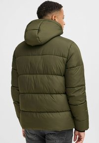 Olivgrønn dunjakke med hette, med quiltet design, glatt tekstur og ribbestrikket kant. Bakfra viser den en strukturert silhuett og sømmer.