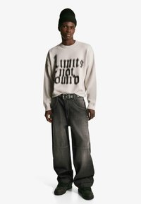 Biały, oversize sweter z czarnym nadrukiem "Limits Not", połączony z luźnymi, szarymi spodniami dżinsowymi i czarnymi sneakersami. Teksturowany splot i mocny materiał.