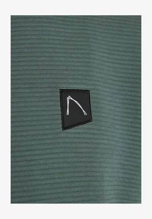 Camisa de tela texturizada de color verde oscuro con un pequeño parche rectangular negro que presenta un diseño gráfico abstracto en blanco en la parte frontal.