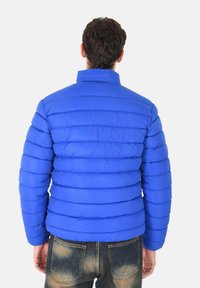 Giacca puffer blu con sezioni trapuntate orizzontali, collo alto e texture liscia, abbinata a jeans in denim sbiadito.