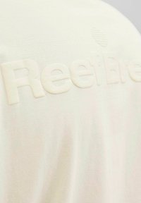 Camiseta de color beige claro con texto en relieve "ReefBreak" y sutiles diseños de texto en el fondo. Tela de algodón suave con un acabado texturizado.