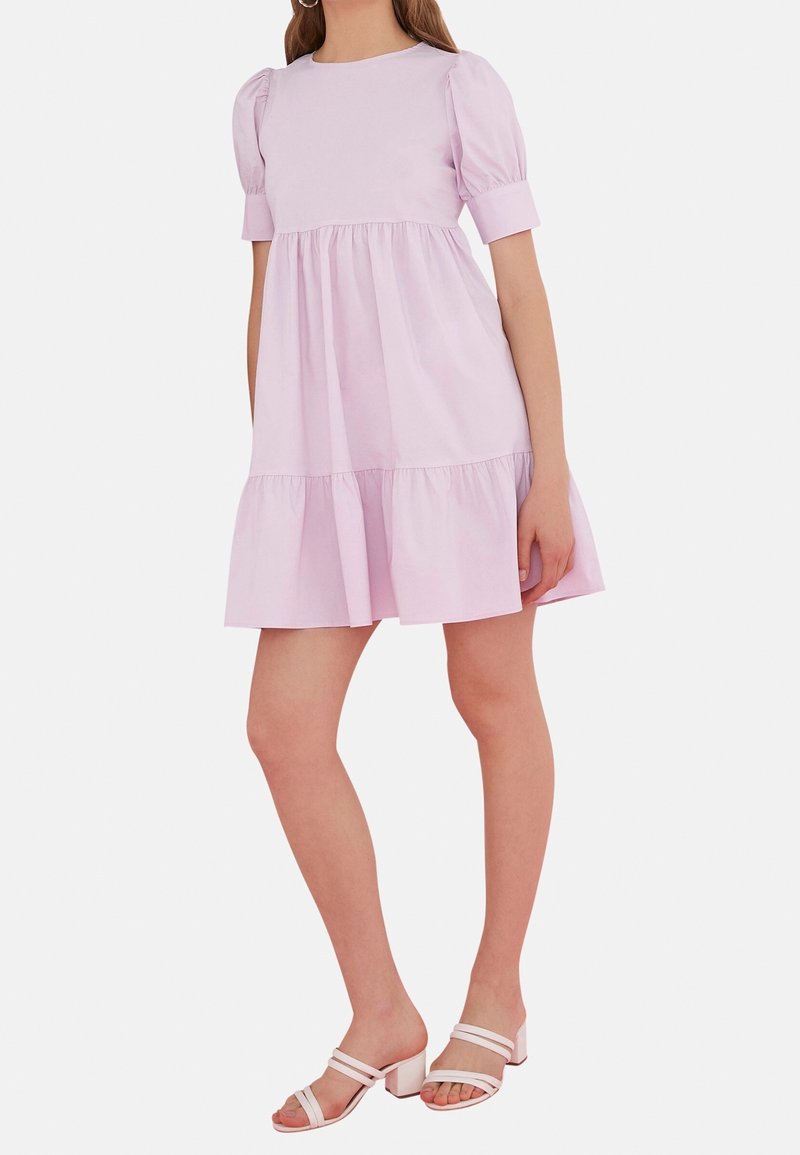 Femme portant une robe courte, violette clair, à manches bouffantes avec une jupe à volants et des sandales à talons blocs ouvertes blanches, se tenant contre un fond blanc.