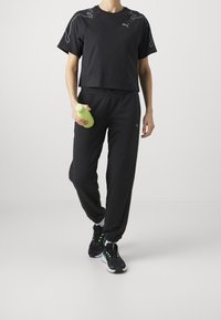 Camiseta deportiva negra cortada con acentos grises, combinada con joggers negros y zapatillas negras. Persona sosteniendo una botella de agua verde.