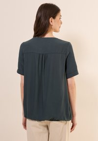 Blusa corta a maniche corte verde scuro con dettaglio plissé sul retro e scollatura rotonda. Realizzata in tessuto leggero e testurizzato.