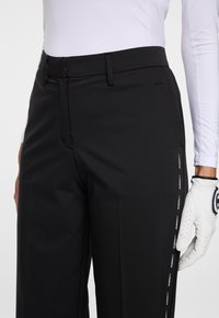 Zwarte op maat gemaakte broek met riemlussen en subtiele zijbranding, gedragen met een wit overhemd met lange mouwen en een witte golfhandschoen aan de rechterhand.