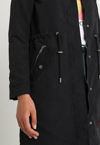 Zwarte parka met een taillekoord, twee zijzakken met ritssluiting en drukknoopsluiting. Gemaakt van een glad, duurzaam materiaal met een losse pasvorm.