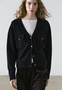 Zwarte cardigan met een V-hals, geribbelde zoom en twee zakken aan de voorkant. Heeft zilveren knoopaccenten. Over een wit shirt gedragen.