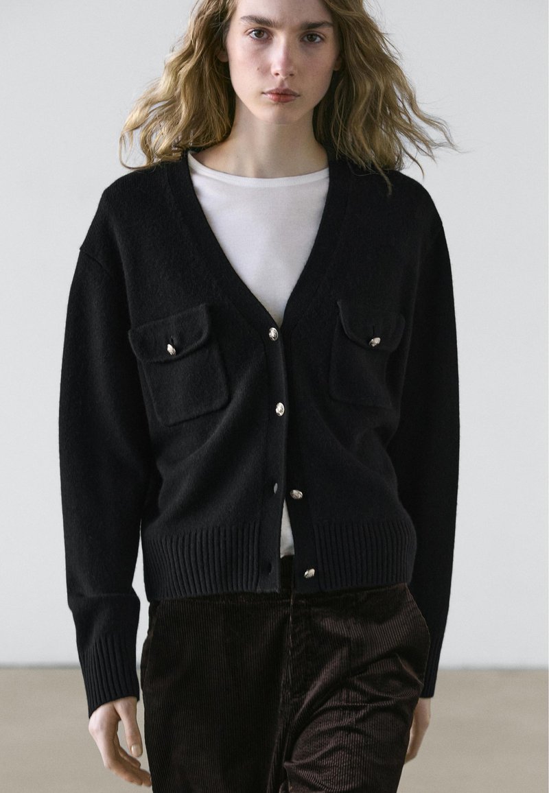 Zwarte cardigan met een V-hals, geribbelde zoom en twee zakken aan de voorkant. Heeft zilveren knoopaccenten. Over een wit shirt gedragen.