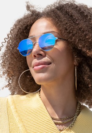Round sunglasses with mirrored blue lenses and metal frames, worn by a person with curly hair, paired with a yellow knitted top and gold jewelry.