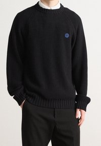 Pull noir en maille avec poignets et ourlet côtoyés, arborant un logo circulaire bleu sur la poitrine. Confectionné en tissu texturé, coupe décontractée.