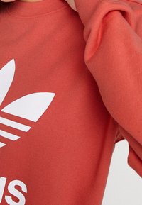 Röd sweatshirt i bomull med ribbad rund halsringning. Har en stor vit Adidas-logotyp och tre ränder på ärmen. Mjuk textur.