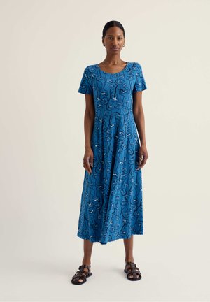 Naine, kes kannab sinist lillemustrit keskmise pikkusega kleiti lühikeste varrukatega, seisab sirgelt hele ühevärvilise tausta ees, jalas mustad sandaalid.