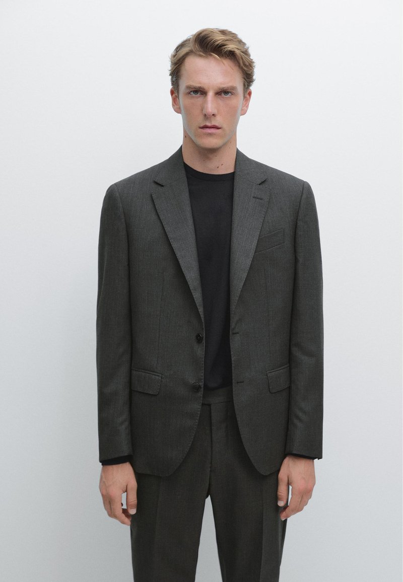 Completo grigio su misura con sottile trama, design monopetto, due bottoni e rever a incrocio, abbinato a una maglietta nera a girocollo.