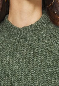 Primo piano di una persona che indossa un maglione a maglia verde testurizzato con colletto a coste e grandi orecchini a cerchio dorati.