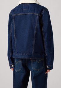 La giacca in denim presenta una finitura blu scuro con colletto in shearling, cuciture a contrasto e una vestibilità classica. Sul retro è visibile un'etichetta del marchio e polsini con bottoni a pressione.