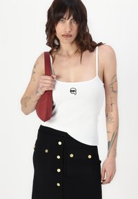 Hvit ribbede tanktop med tynne stropper som har en liten svart logo, kombinert med et svart skjørt prydet med gullknapper, og en rød håndveske.