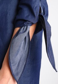 Tummansininen farkkupaita, jossa on solmittu yksityiskohta hihassa, ja läpinäkyvä, kontrastina oleva vaaleansininen sisäkerros, joka tuo lisärakennetta ja visuaalista mielenkiintoa.