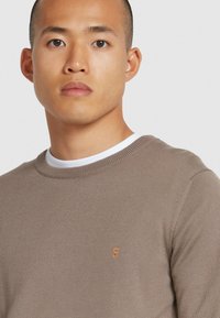 Brązowy sweter dziany z okrągłym dekoltem, z pomarańczowym haftowanym logo na klatce piersiowej i teksturowanym wykończeniem.