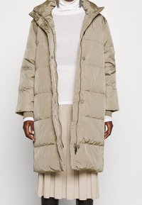 Lys beige pufferjakke med høj krave, synlige syninger, frontlynlås og knaplukninger. Blødt, vandtæt materiale med quiltet design.