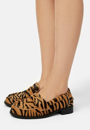 Mocassins plats en fourrure tan avec des rayures noires de tigre. Design à bout rond, petit talon carré, et détails de couture visibles sur le dessus.