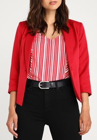 Roter Blazer über einem rot-weiß gestreiften Oberteil, kombiniert mit schwarzen hochgeschnittenen Hosen und einem schwarzen Gürtel mit einer silbernen Schnalle.