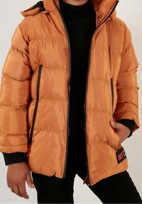 Chaqueta acolchada naranja con cremallera frontal, detalles en negro, capucha y dos bolsillos laterales. Presenta un parche con logo y una textura brillante y suave.